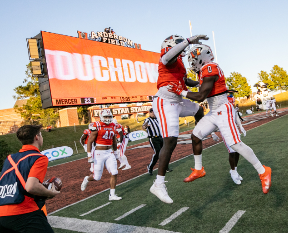 Two Mercer football players wearing orange jersey chest bump in the air in the end zone of a football field. The score board in the back says, 