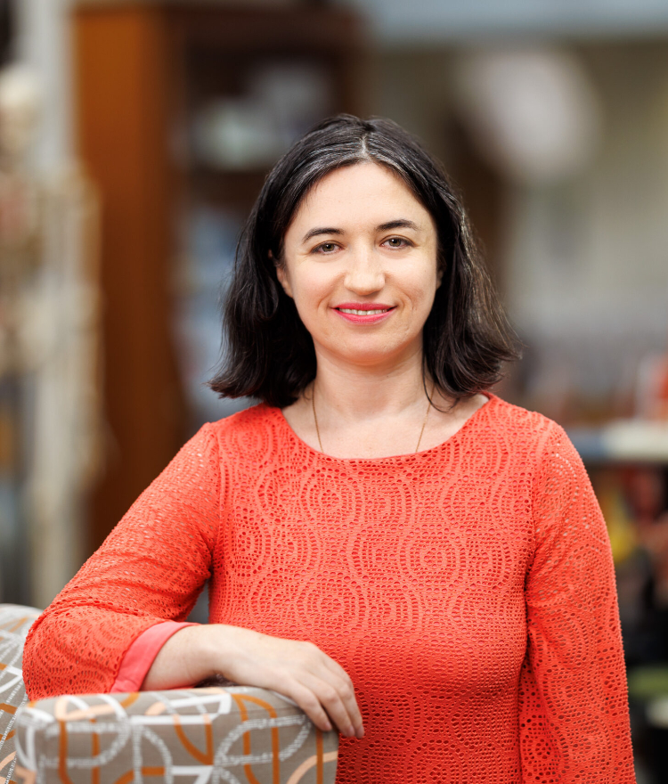 Dr. Ilana Chefetz Menaker is smiling while wearing an orange shirt. Dr. Menaker is standing next to a modern chair, with anatomical models in the background, suggesting an educational or medical setting.