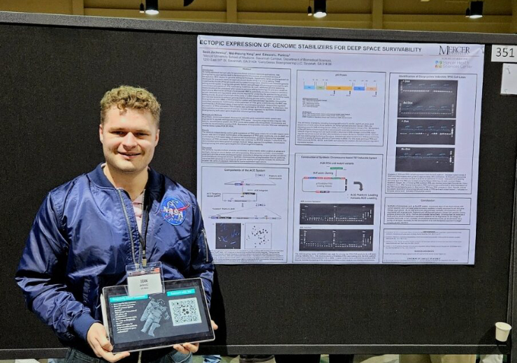 A Mercer student wearing a blue jacket with a NASA patch holds iPad that displays a space suit. He is standing next to a poster titled: Ectopic Expression of Genome Stabilizers for Deep Space Survivability.