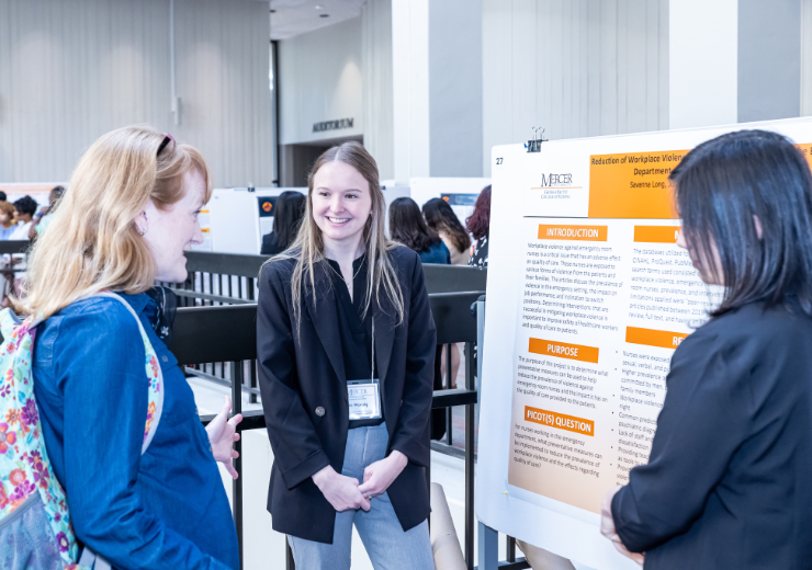 Two Mercer students wearing business attire stand on either side of their research poster as they interact with a woman facing the poster.