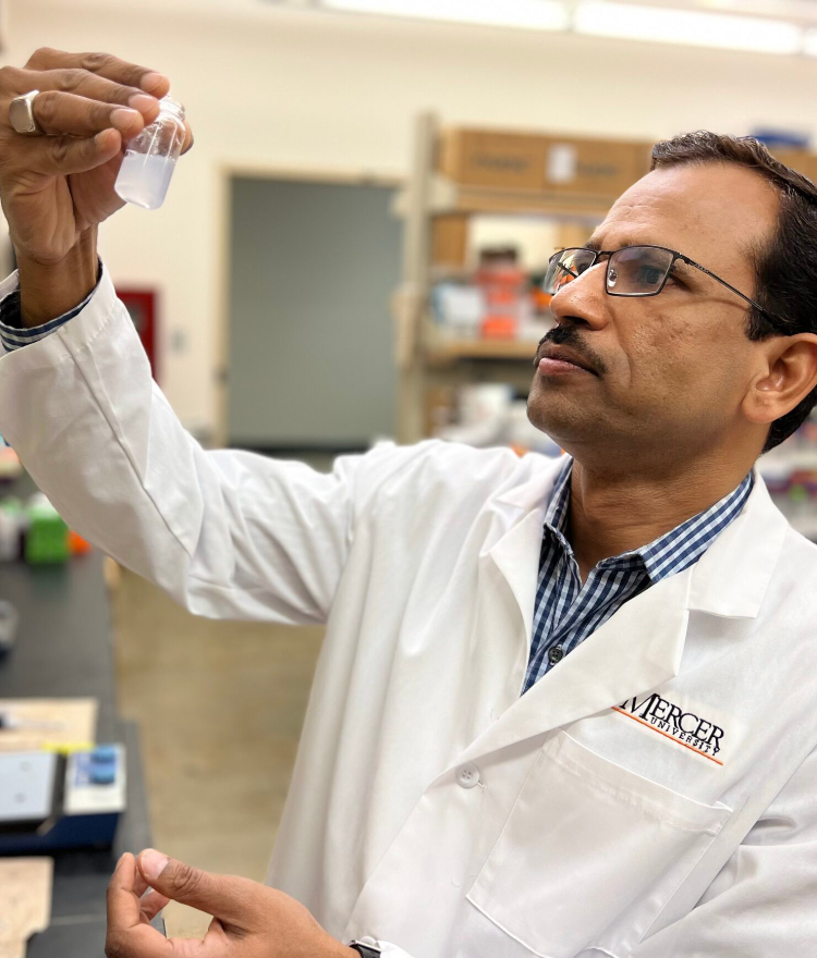 Dr. Mahavir Chougule wearing a white lab coat and holding up a vial of liquid in a laboratory.