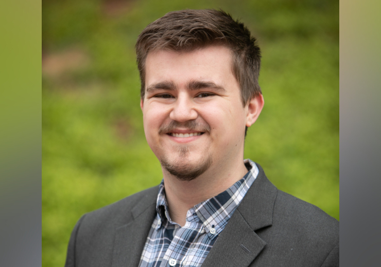 Headshot of Caden Hamrick wearing a plaid, button up shirt and gray blazer in an outdoor setting.