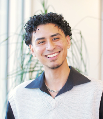 Portrait of a smiling Daniel Alejandro Mendoza with curly hair, wearing a gray sweater over a collared shirt, standing in a bright room with plants in the background.