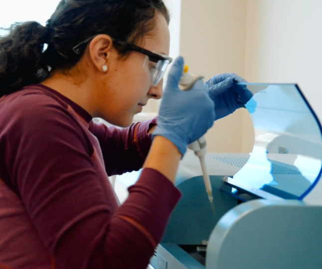 A Mercer student wearing protective eyewear and gloves uses leans over an open piece of lab equipment with a dropper.