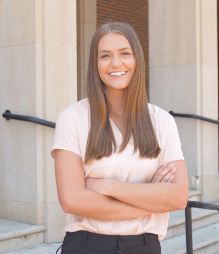 Elizabeth Merchant smiling and standing with crossed arms in front of a building entrance.