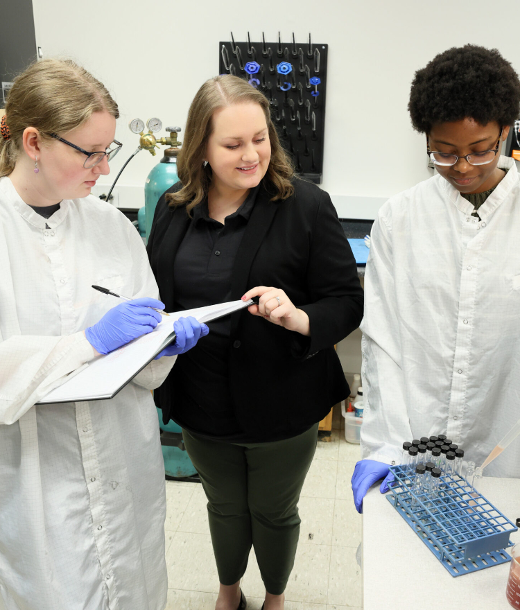 Dr. Sarah Bauer, wearing black business casual attire, and two Mercer students wearing lab coats working in a lab.