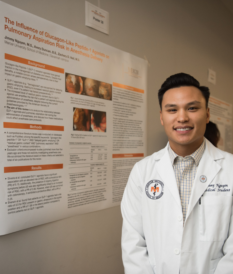 A Mercer School of Medicine student wearing a white lab coat stands beside his research poster titled, 