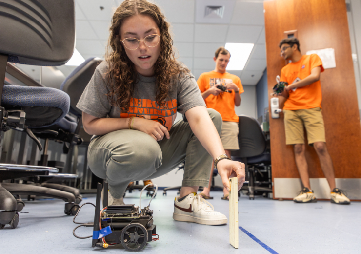 A Mercer University student crouches on the ground holding a small panel of wood in front of a small, wheeled robot.