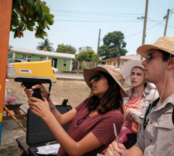 Three individuals use a pXRF device to test for lead on a metal bar with peeling orange paint outdoors. The person to the left operates the device as another takes notes, and a third observes. All are wearing sun hats.