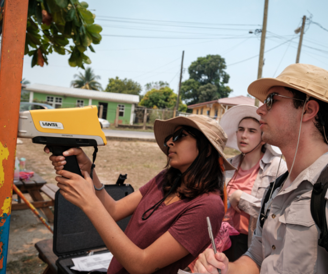 Three individuals use a pXRF device to test for lead on a metal bar with peeling orange paint outdoors. The person to the left operates the device as another takes notes, and a third observes. All are wearing sun hats.