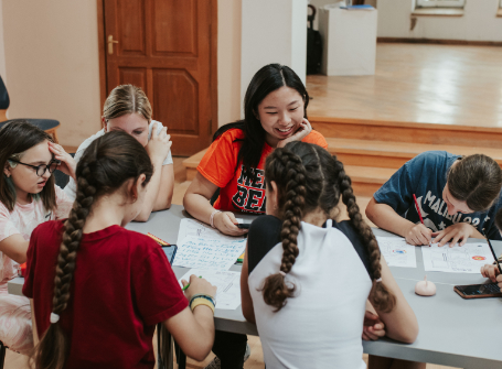 A student wearing an orange Mercer T-shirt sits at a table with children writing on papers.