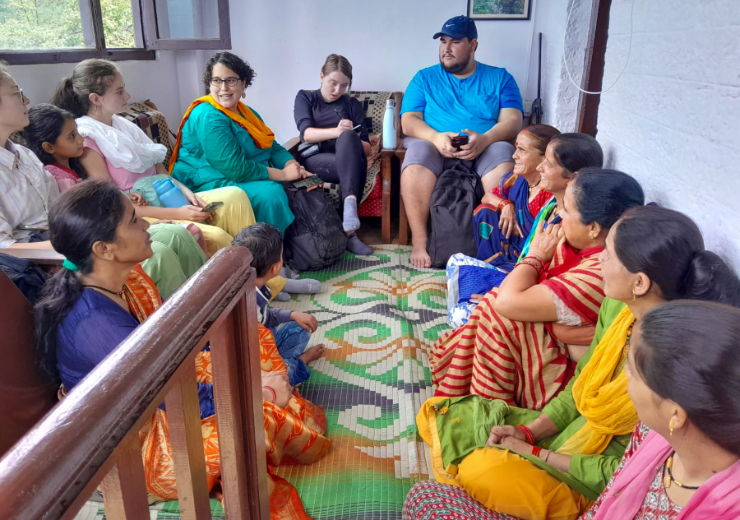 Mercer students, a professor, and Uttarakhand residents sit in a circle in a room of a home. The Uttarakhand residents are wearing brightly colored traditional India clothing and sitting on the floor. The Mercer students and professor sit on a couch and chair. One student is taking notes.