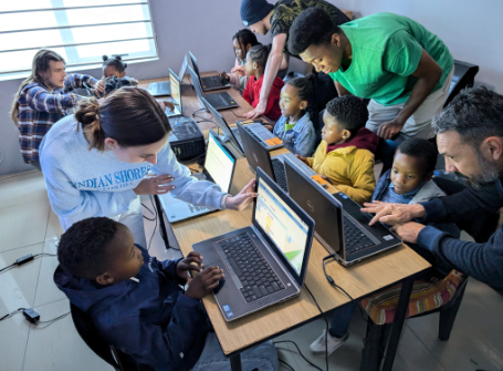 Eight children work on computers at a desk; four children are on either side. Four Mercer students lean over the children helping them.