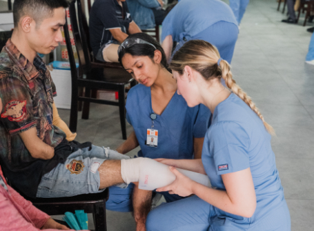 Two Mercer students wearing blue scrubs fit a prosthetic leg on a Vietnamese man sitting in a chair.