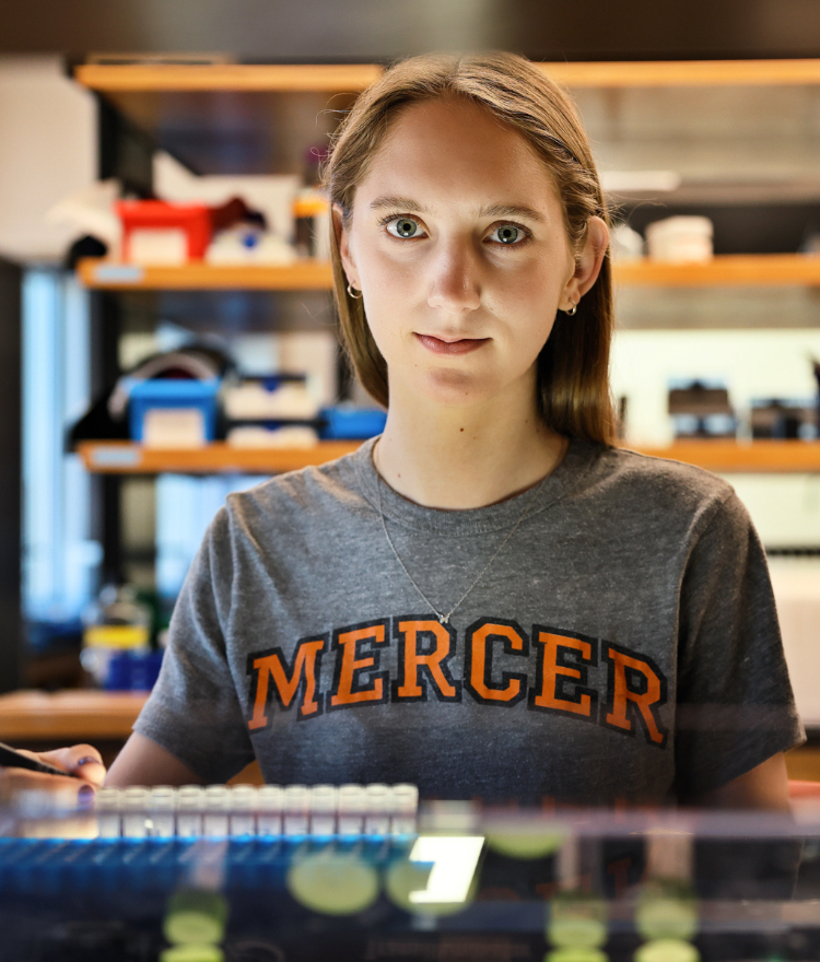 Sarah Boyer wearing a Mercer shirt looks at the camera. A lab is in the background.