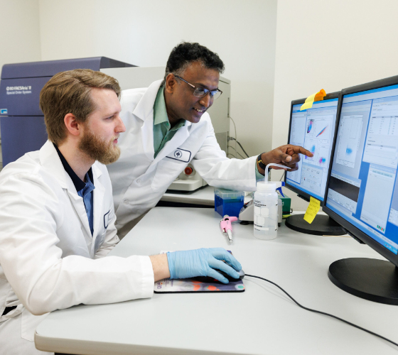 Dr. Raghavan Chinnadurai and biomedical sciences student working in a lab. The student controls a computer mouse, and Dr. Chinnadurai points at the computer monitor.