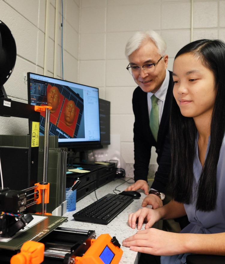 A Mercer professor is seen over the shoulder of a student as they watch a 3D printer. Two molds of faces are see on a computer screen in the background.