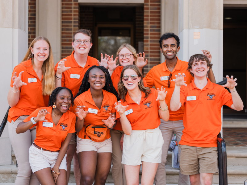 Eight diverse individuals wearing orange T-shirts and khaki shorts make bear claws with their hands.