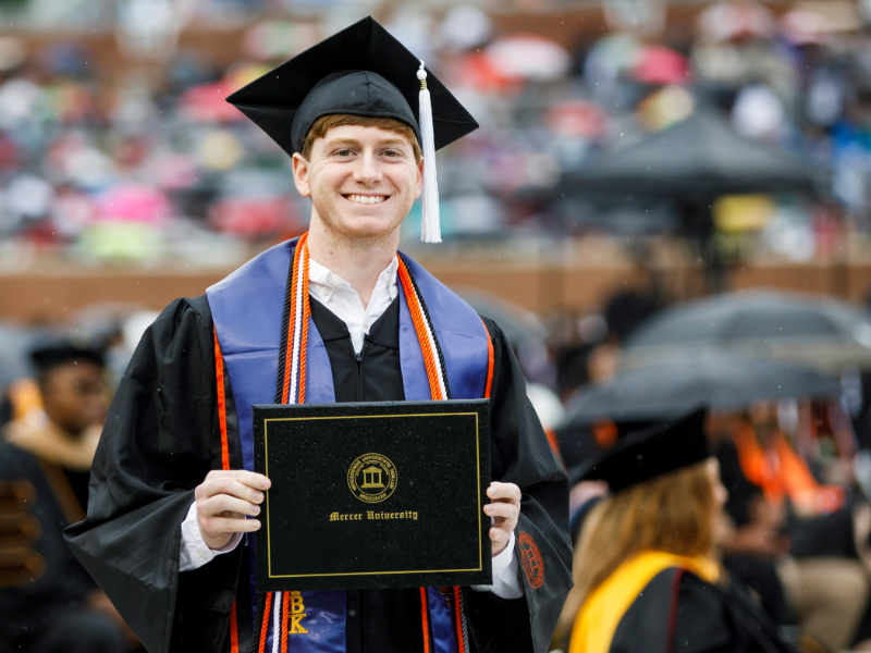 An individual wearing graduation attire holds a Mercer University diploma.