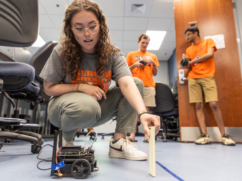 A Mercer University student crouches on the ground holding a small panel of wood in front of a small, wheeled robot.