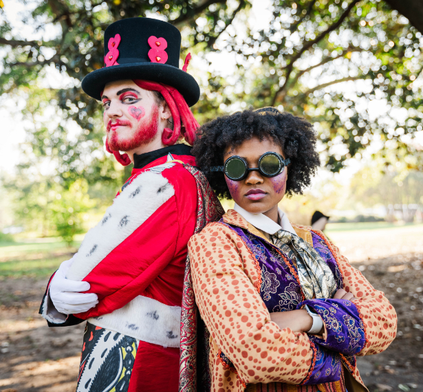 Two people wearing brightly colored costumes in a steam punk style stand back-to-back with their arms crossed.