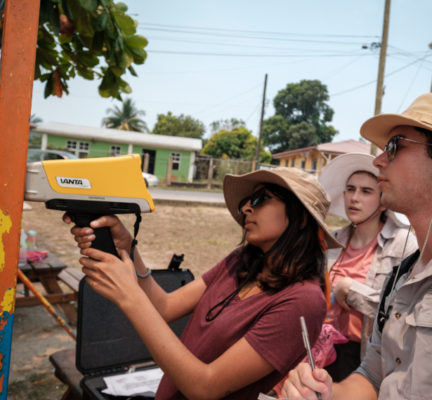 Three individuals use a pXRF device to test for lead on a metal bar with peeling orange paint outdoors. The person to the left operates the device as another takes notes, and a third observes. All are wearing sun hats.