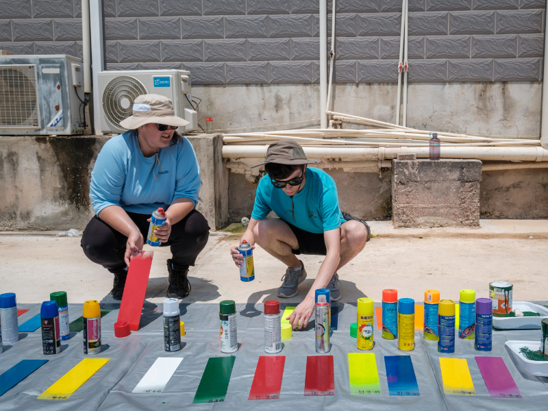 Two individuals, wearing caps and casual attire, hold cans of spray paint while more cans and painted color swatches are laid out on a tarp on the ground in front of them.