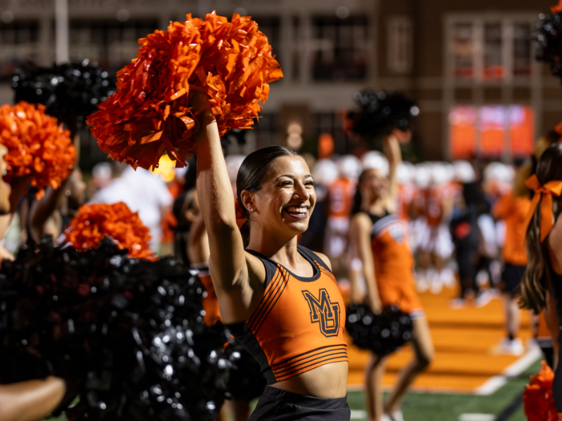 An individual wearing an orange Mercer cheerleading uniform waves a pom pom during a football game.