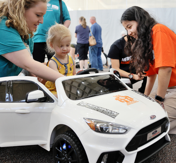 A Mercer student wearing an orange T-shirt and a woman wearing a teal T-shirt interact with a toddler in a white, modified battery-powered toy car.