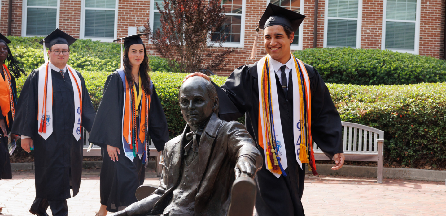 A student in graduate cap and gown rubs the head of the Jesse Mercer statue.