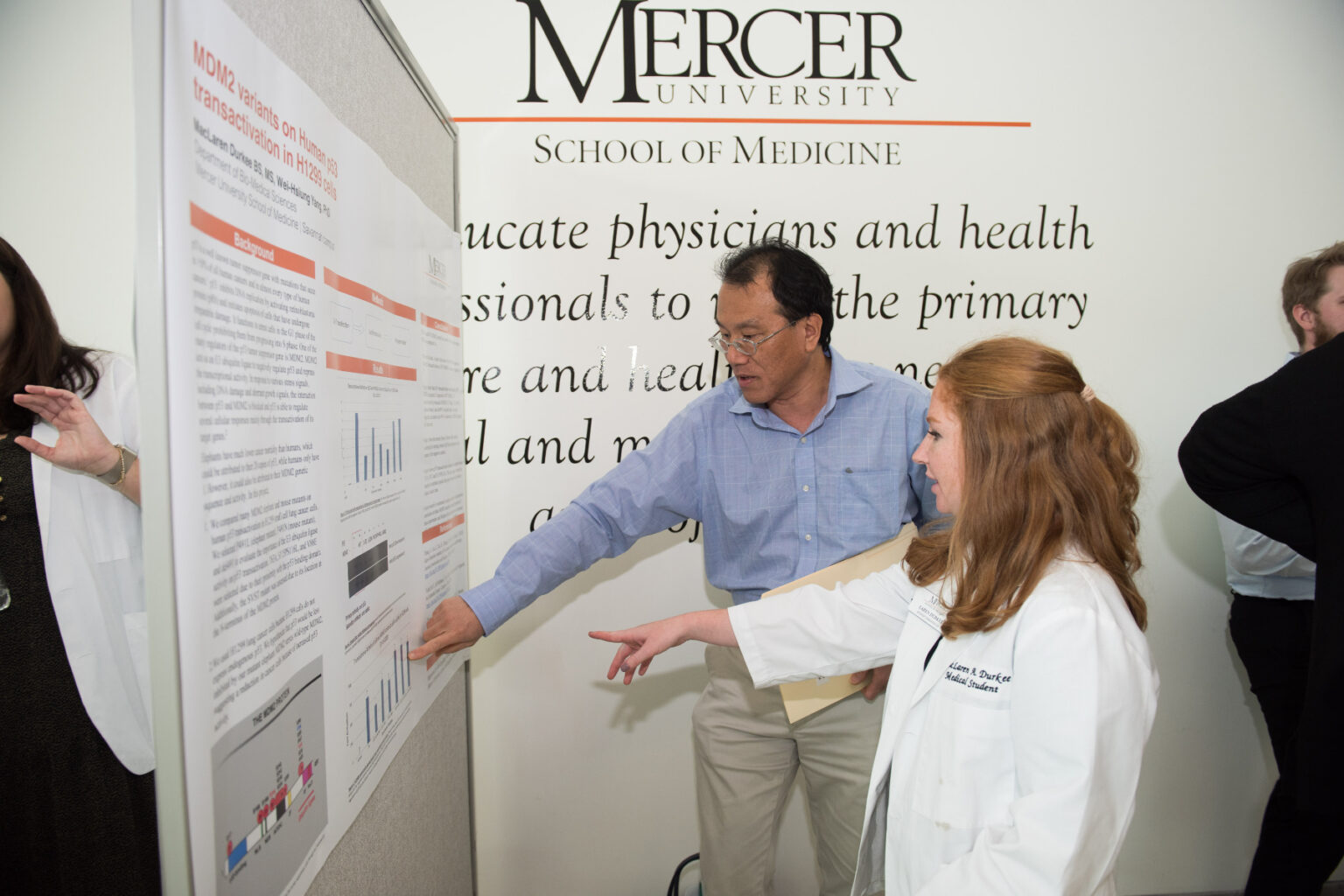 A Mercer medical student in a lab coat points at a poster next to a man also pointing at the poster.