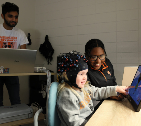 Parth Patel stands at a computer, while Lynzi Holland sits at a table with a child wearing a cap with the fNIRS technology.